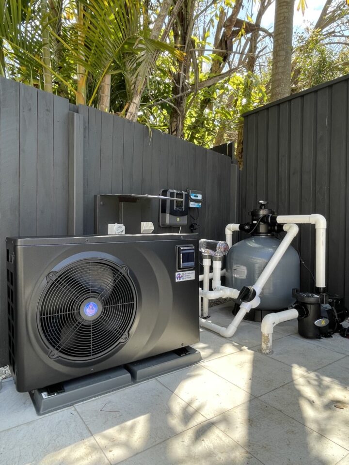 A backyard setup featuring a black pool heat pump and a sand filter system with white PVC piping. The equipment is positioned on a tiled surface with a dark wooden fence and lush foliage in the background. Bright sunlight illuminates the scene. | AES Pool Heating Solutions